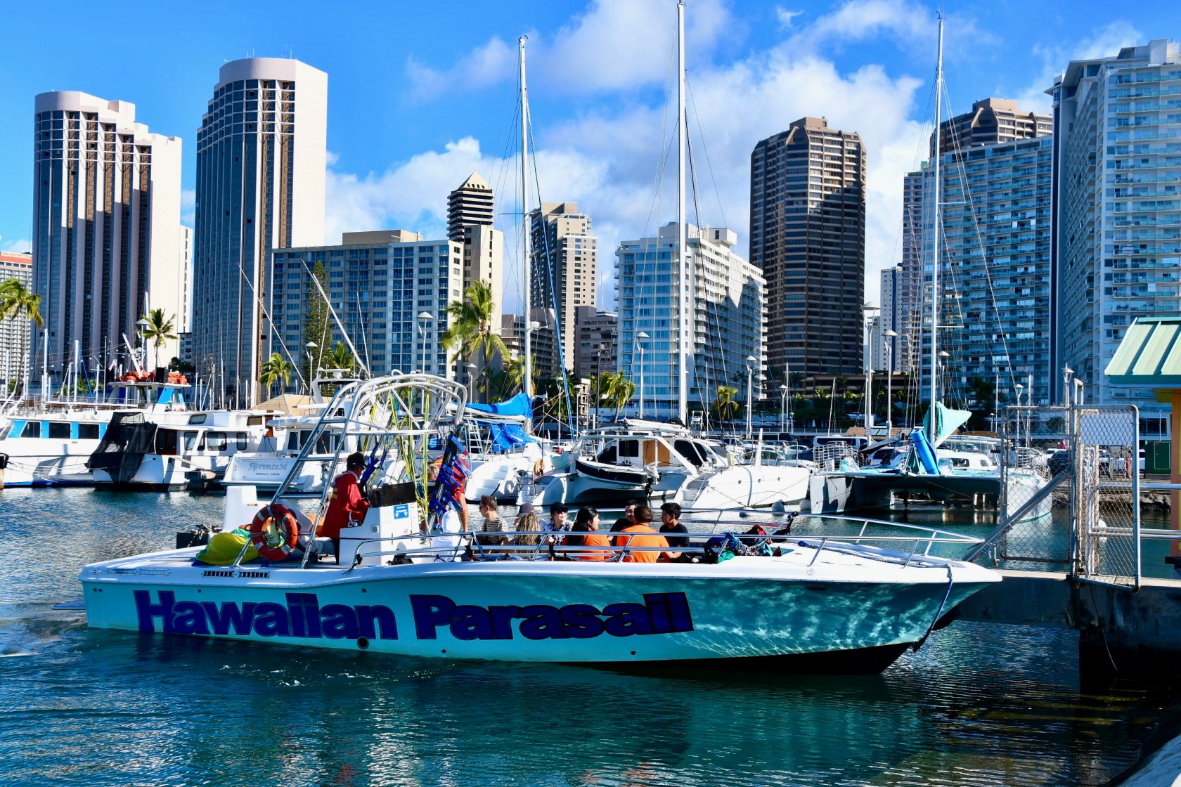 Boat labeled 'Hawaiian Parasail' docked in marina with skyscrapers in background.